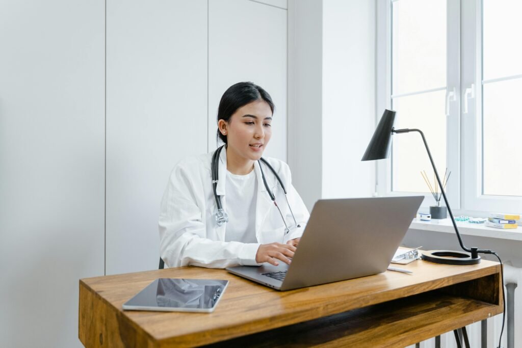 A female doctor in a white coat uses a laptop for an online consultation from her office.
